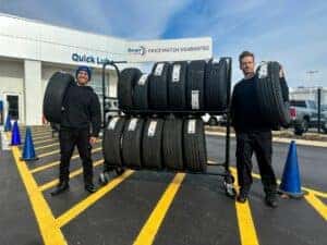 Two guys holding tires next to a tire rack
