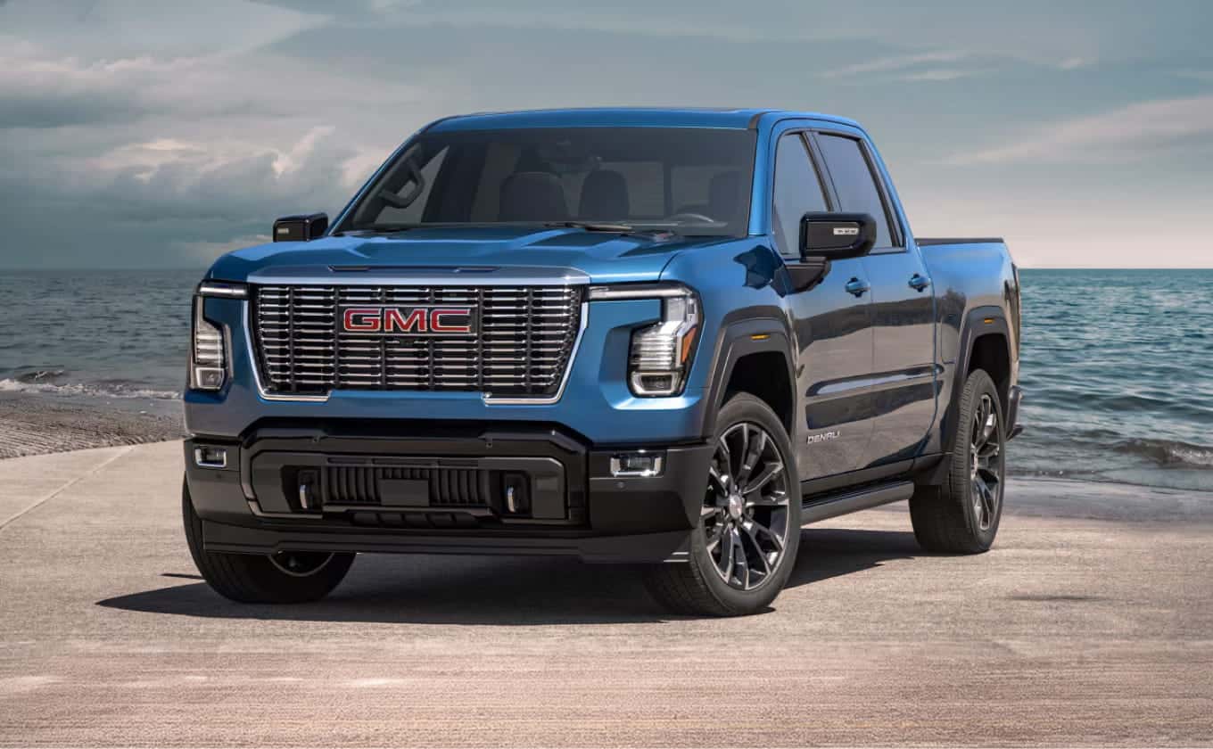 Front three-quarter view of a gmc blue pickup truck with a chrome grille parked on a paved surface beside the ocean.