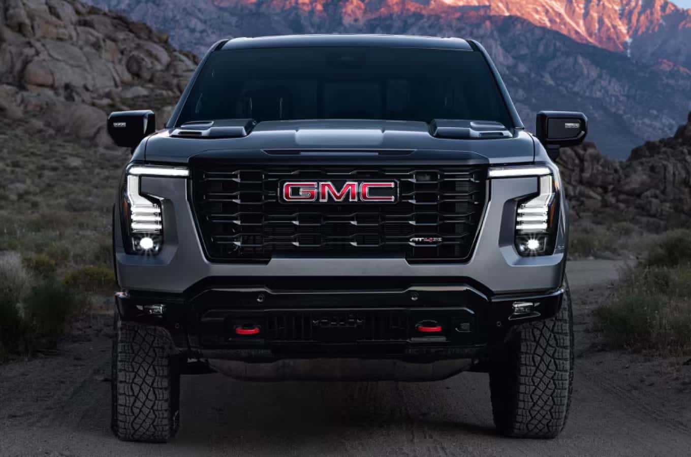 Front view of a gmc dark gray pickup truck with illuminated LED headlights and rugged tires against a rocky mountain backdrop.