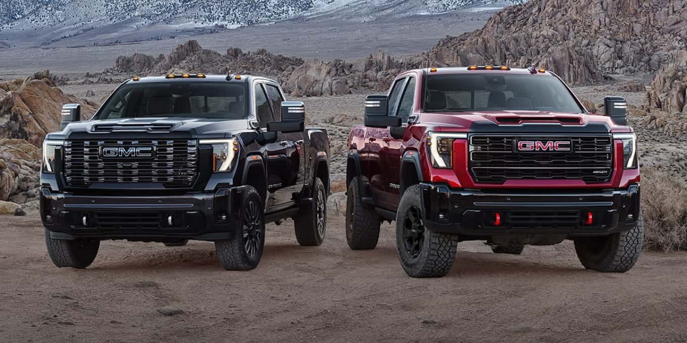 Two gmc heavy-duty pickup trucks in black and red parked side by side on rugged terrain with rocky mountains in the background.