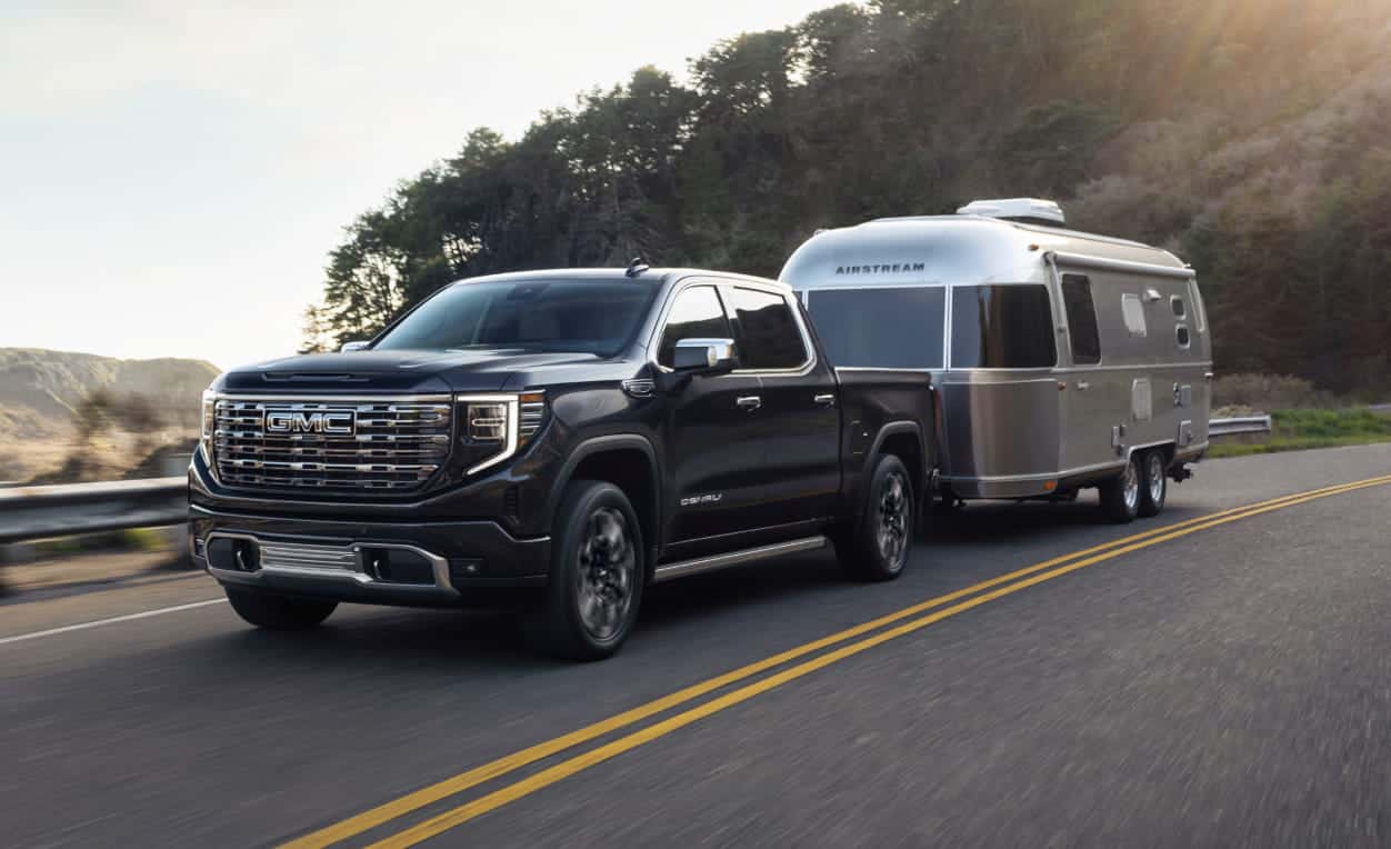 A black 2026 GMC Sierra 1500 truck tows a silver Airstream trailer along a scenic, winding road with green hills and sunlight in the background.