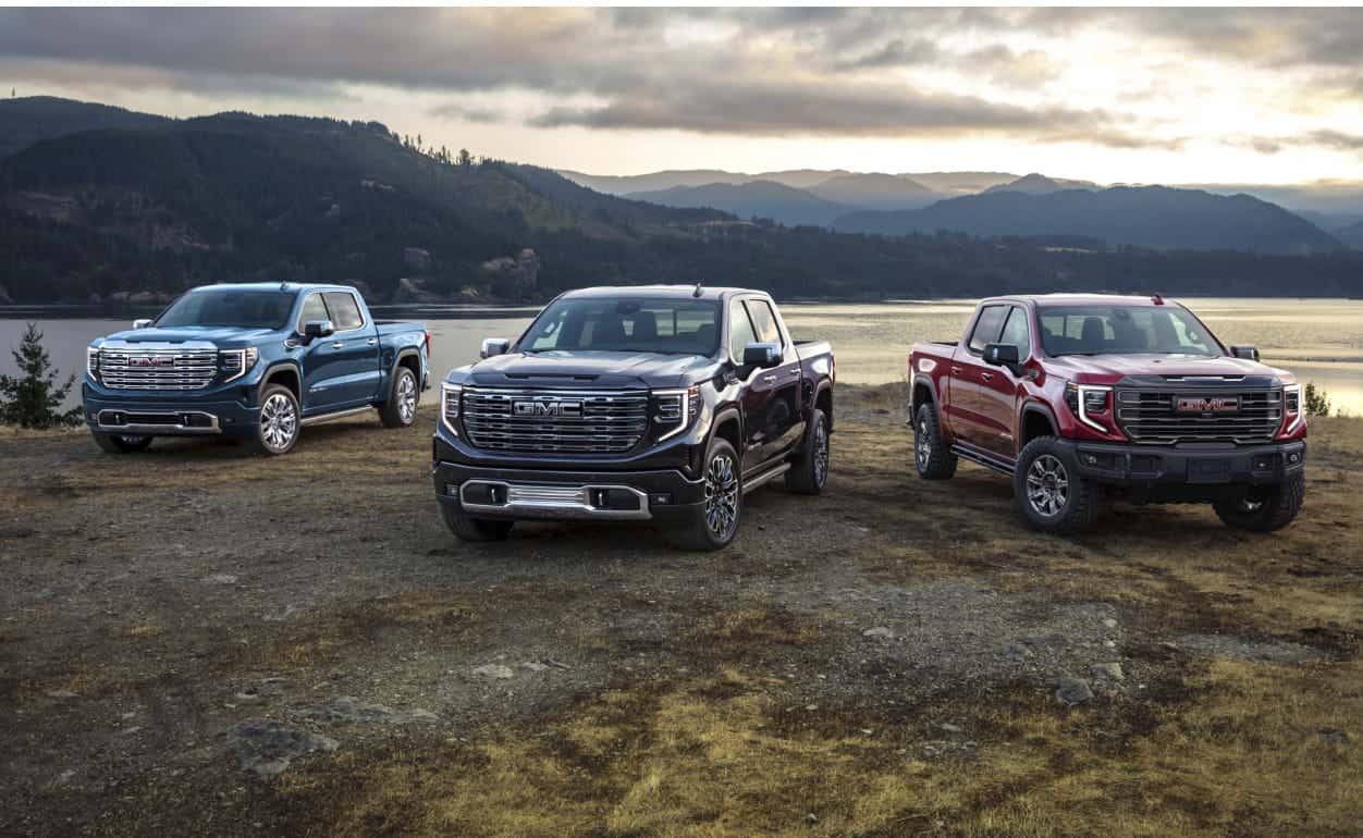 Three 2026 GMC Sierra 1500 trucks are parked on a rocky lakeside with mountains in the background under a cloudy sky.