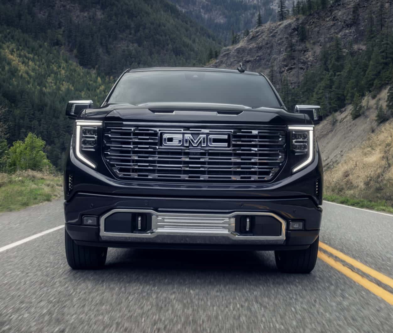 Front view of a black 2026 GMC Sierra 1500 truck on a road amidst forested mountains.