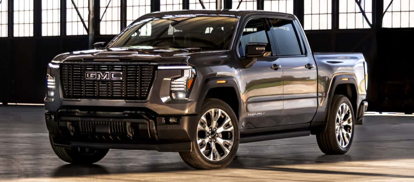 Front three-quarter view of a gmc dark gray pickup truck with a large chrome grille and polished wheels inside an industrial warehouse.