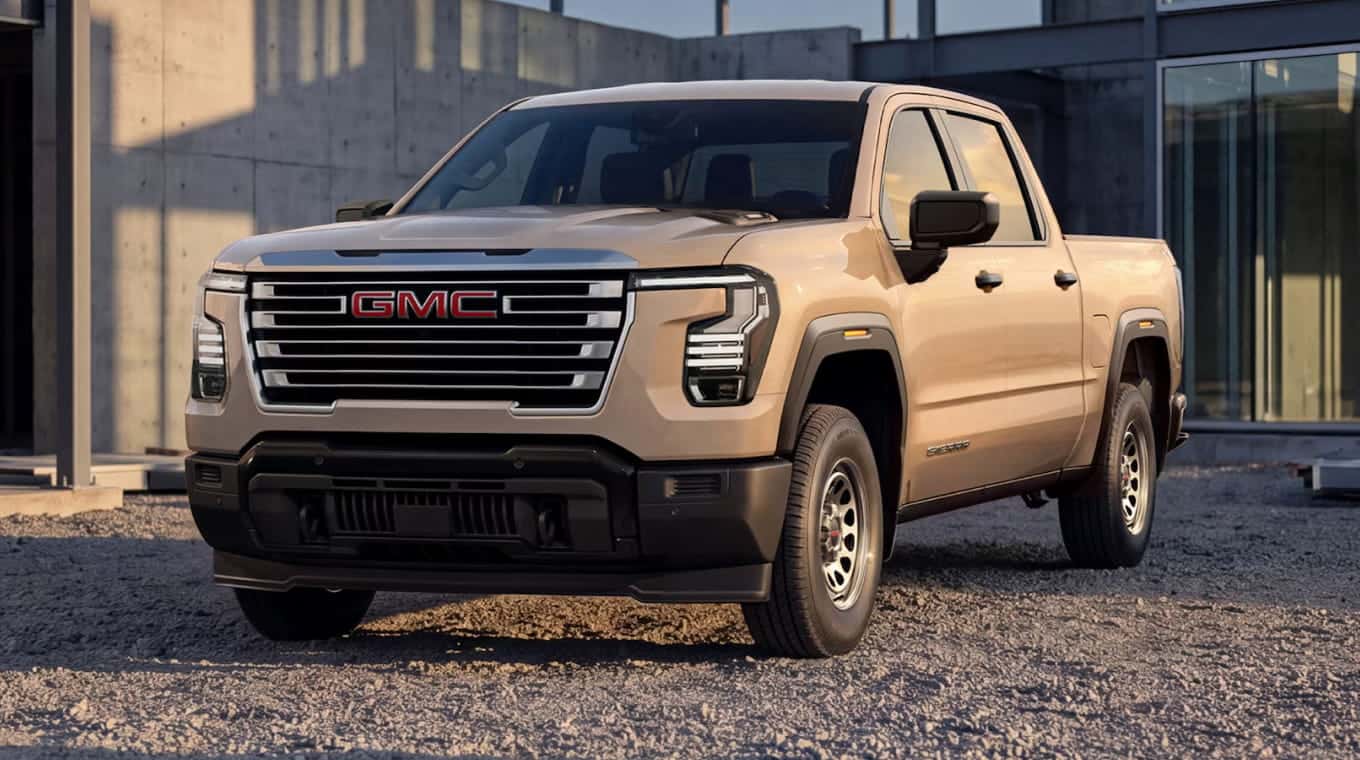 Front three-quarter view of a gmc beige pickup truck with a chrome grille parked on gravel outside a modern building.