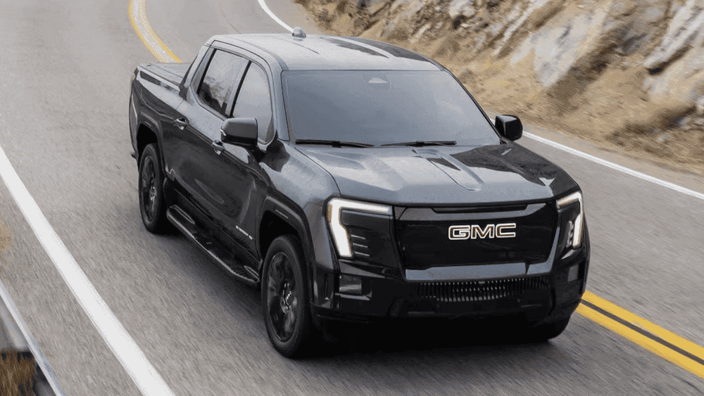 2026 GMC Sierra EV near Ruth NC in gloss black with bold GMC grille lettering, LED signature lighting, and blacked-out alloy wheels driving on a mountain highway
