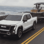 2026 GMC Sierra EV near Ruth NC in white towing a sport boat along a coastal highway with bold GMC grille lettering, LED headlights, and AT4 off-road badging