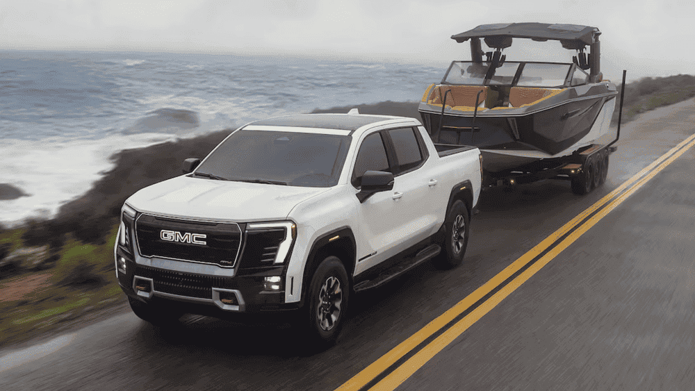 2026 GMC Sierra EV near Ruth NC in white towing a sport boat along a coastal highway with bold GMC grille lettering, LED headlights, and AT4 off-road badging