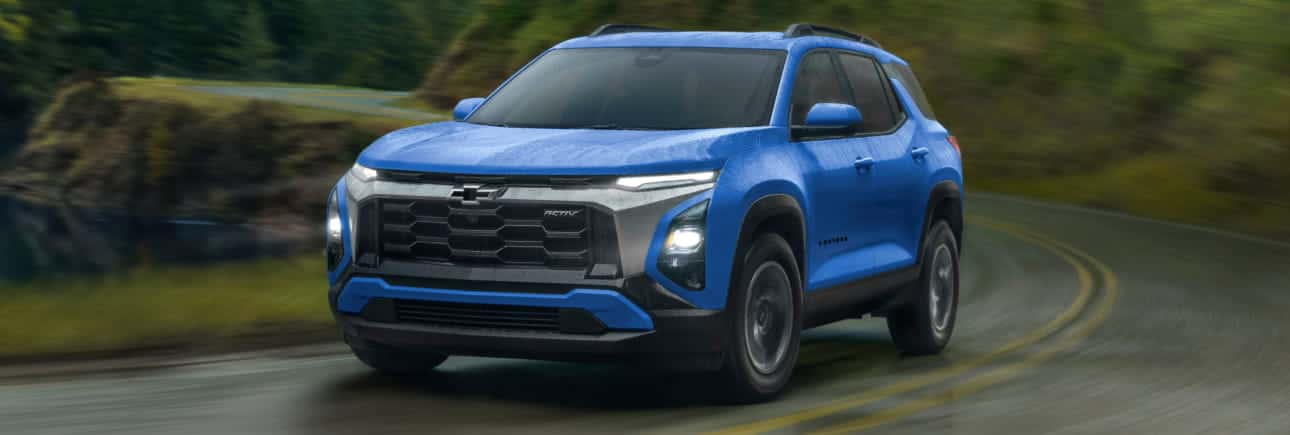 A blue Chevrolet Equinox ACTIV SUV driving along a winding road surrounded by lush greenery, with its headlights illuminated.