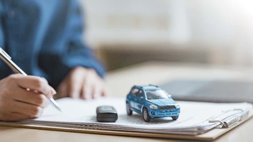A person signing a vehicle contract with a key fob and blue, toy SUV on top of the paperwork.