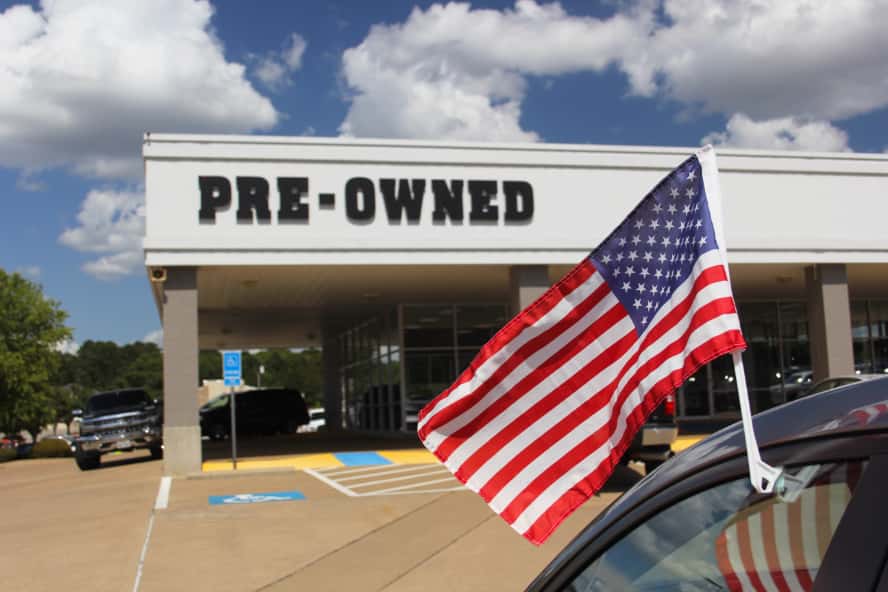 A pre-owned vehicle dealership with an American flag in the foreground attached to a car's window.