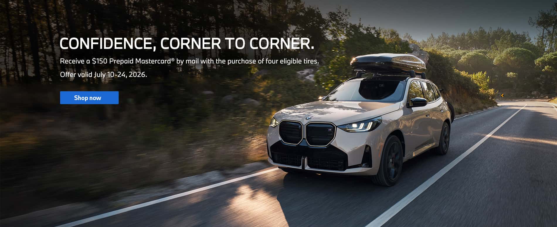 A front, three quarter view of white BMW X3 is driving on a tree-lined, paved road. A BMW roof box is attached to the top of the vehicle for extra storage.
