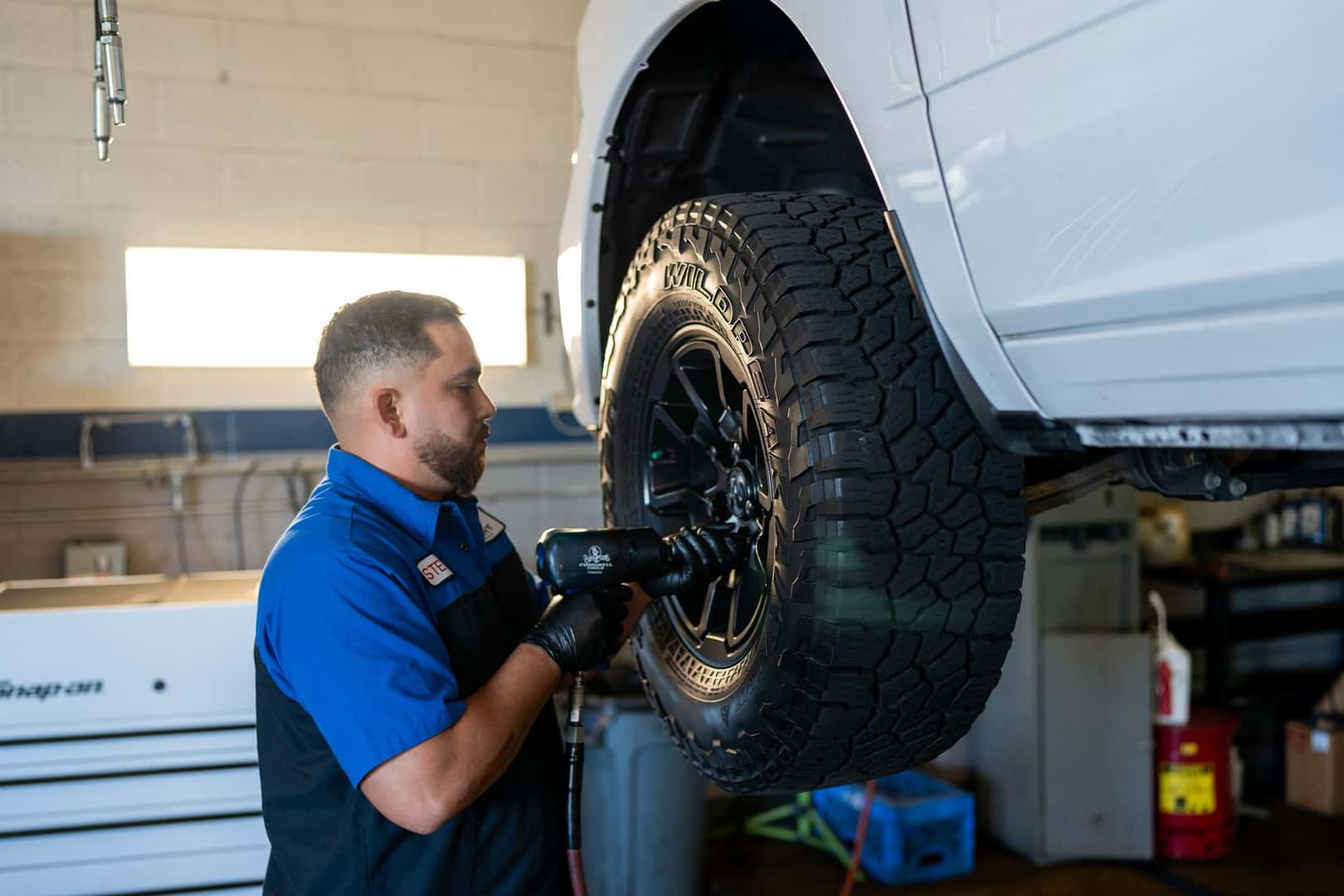 A technician at Knight Claremont Chrysler Dodge Jeep Ram using an impact wrench on an all-terrain tire and black wheel of a white pickup on a lift