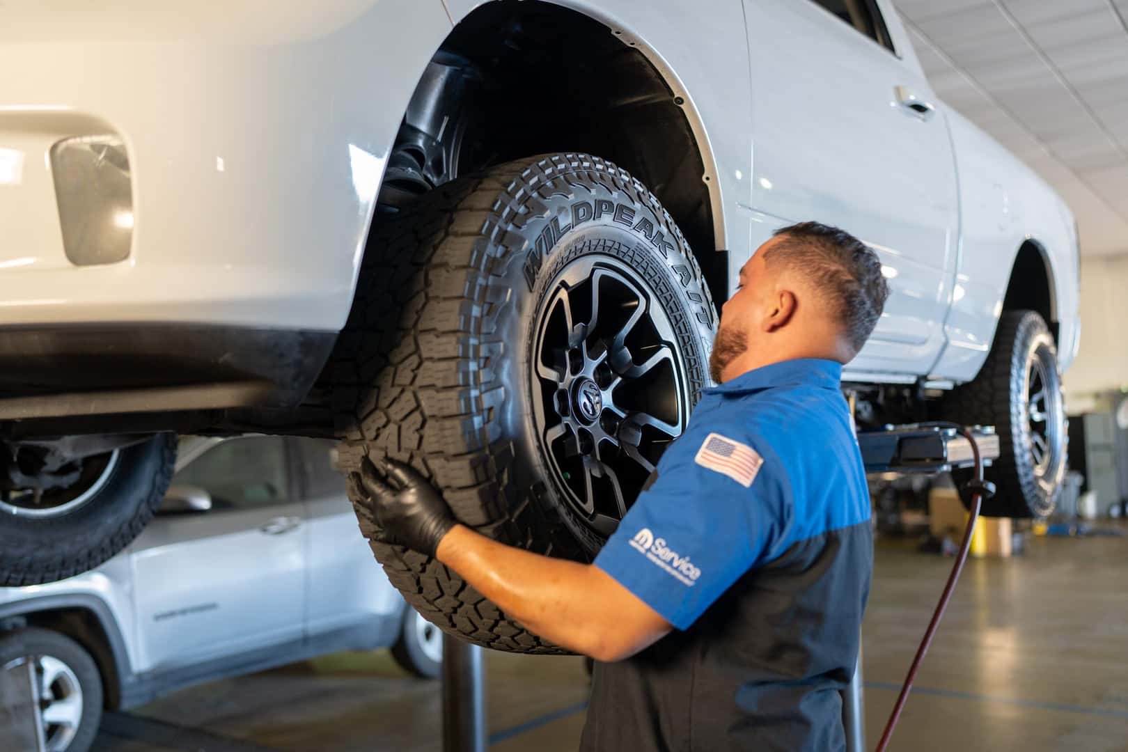 A technician at Knight Claremont Chrysler Dodge Jeep Ram fitting an all-terrain tire and wheel to a white pickup on a lift in the service shop