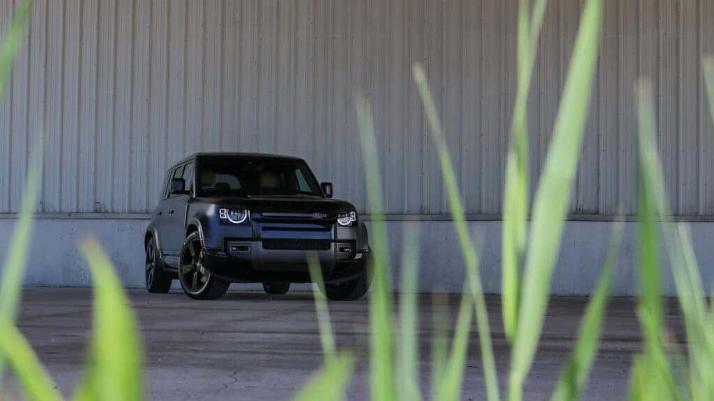 Front view of a 2025 Land Rover Defender parked outdoors, showcasing its bold grille, rugged body lines, and elevated stance, with off-road terrain in the background.
