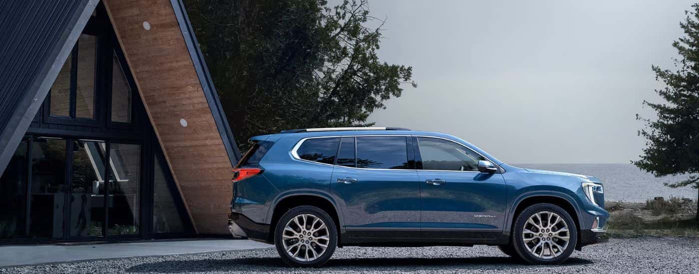 Blue 2026 GMC Acadia Denali parked in front of a lakeside cabin.