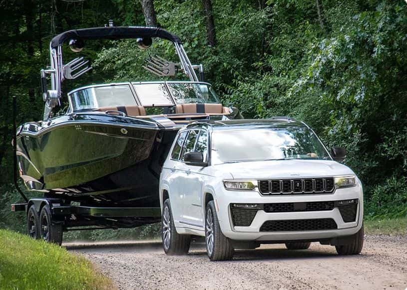 White 2026 Jeep Grand Cherokee L towing a large wakeboard boat on a trailer along a gravel road through trees