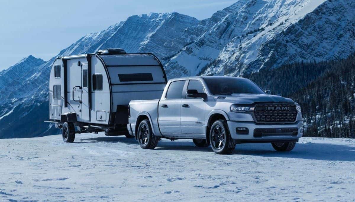 White 2026 Ram 1500 Big Horn 4x4 Crew Cab towing a travel trailer across snow with mountains behind