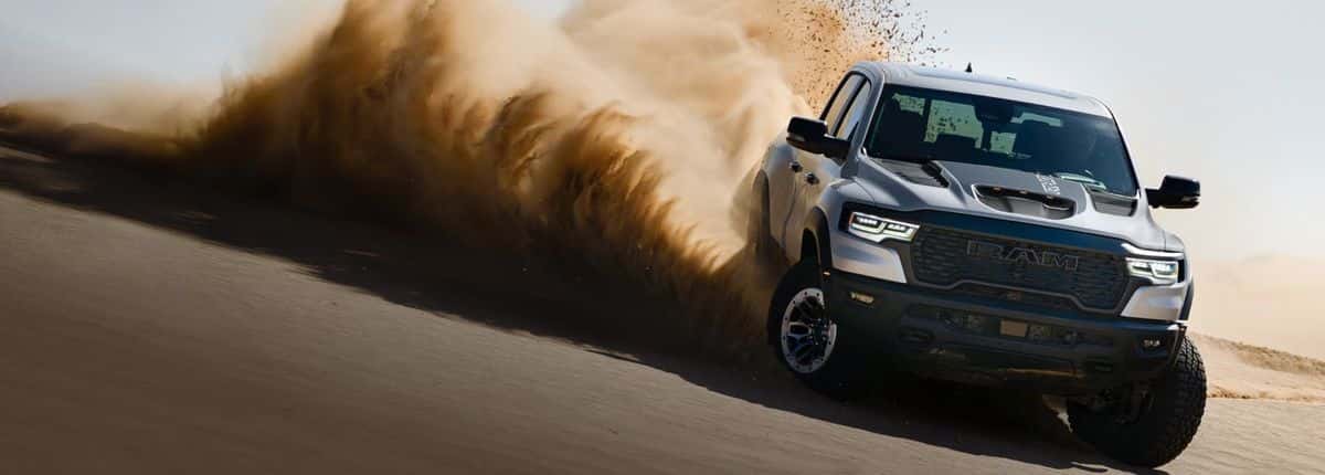 Silver 2026 Ram 1500 RHO 4x4 Crew Cab charging down a sand dune with dust behind it