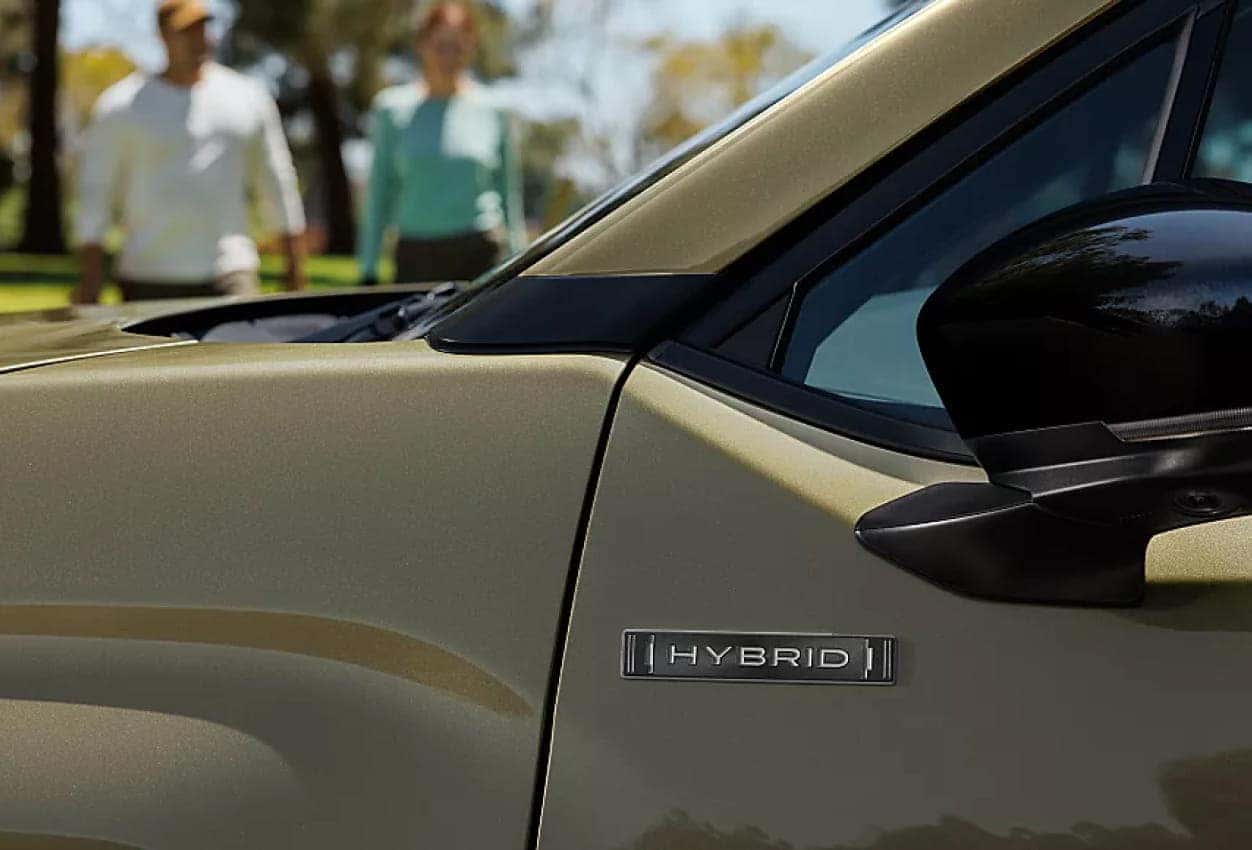 Close-up of a 2026 Subaru Forester vehicle's side with a badge reading Hybrid, and two people walking in the blurred background. Sunny, outdoors setting.