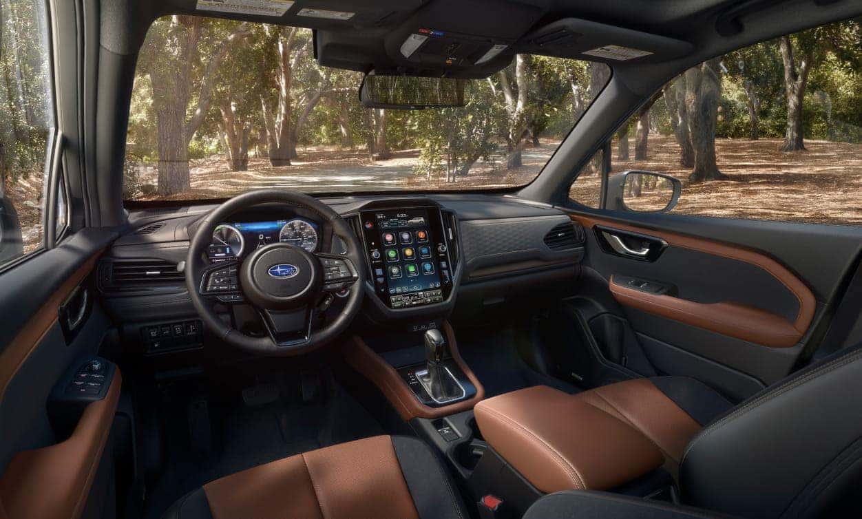 The interior of a 2026 Subaru Forester features a sleek dashboard with a large touchscreen, a leather-wrapped steering wheel, and brown and black leather seats, set in a wooded area.