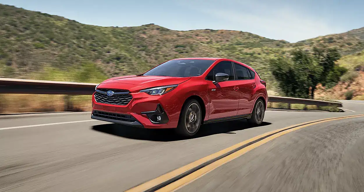 Red 2026 Subaru Impreza driving along a winding mountain road with rolling hills and scenic countryside in the background