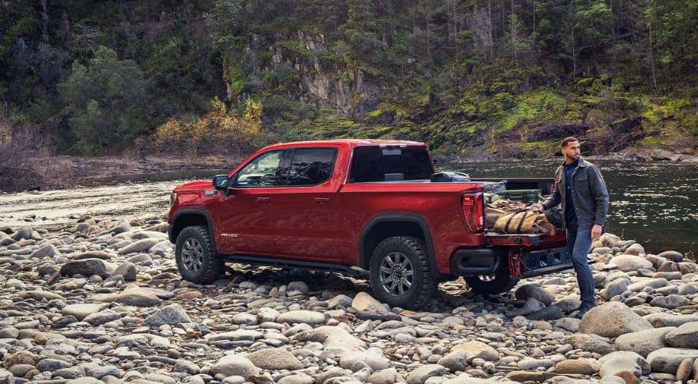 A person loading the bed of a red 2026 GMC Sierra 1500 AT4X on a rocky riverside.