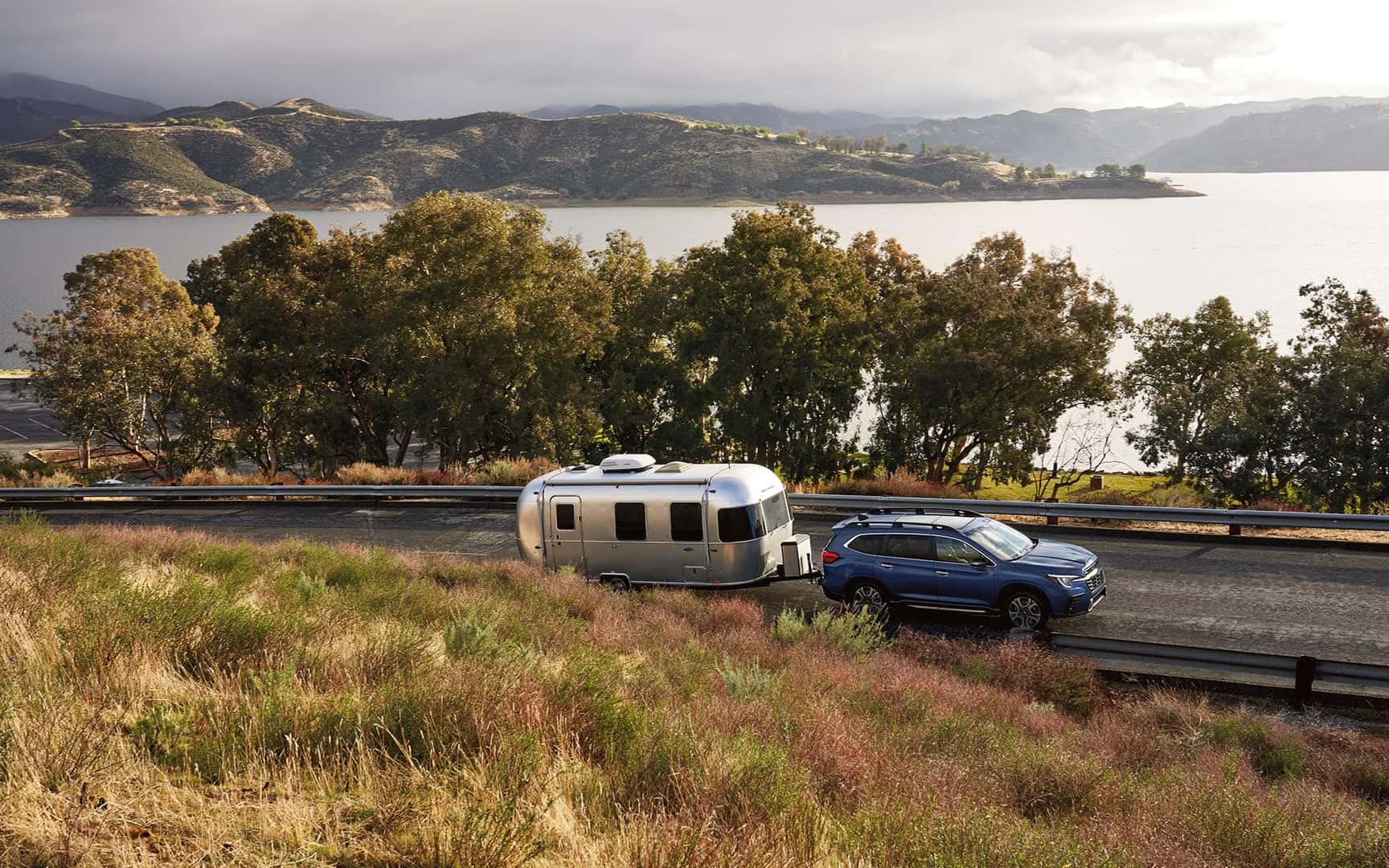 Profile view of Ascent towing Airstream trailer uphill with trees and lake in background