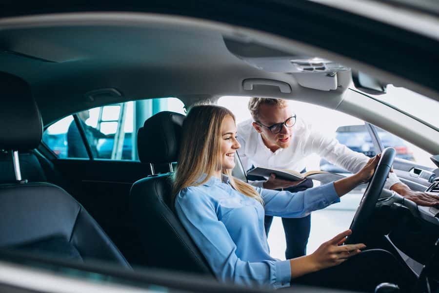 A woman sits behind the wheel of a car while a salesperson leans in to show her the controls