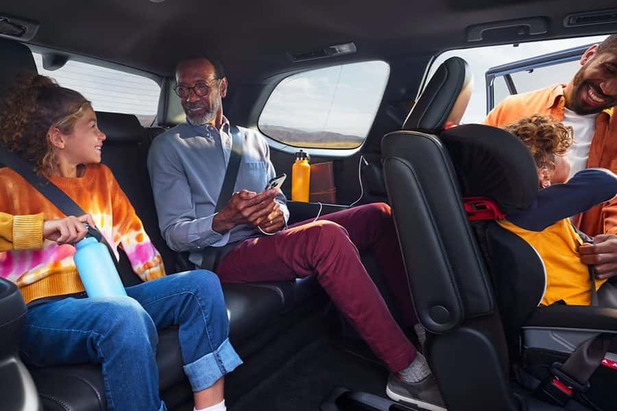 A family sits in the back rows of an SUV while a parent buckles a child into a car seat
