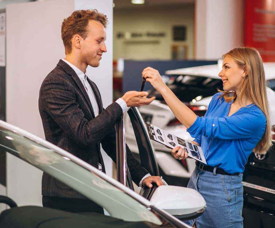 A salesperson hands car keys to a customer beside a car in a showroom