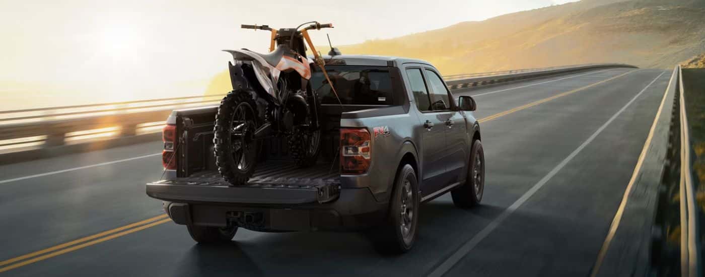Rear view of a grey 2026 Ford Maverick FX4 with a dirt bike in the bed driving on a paved road.