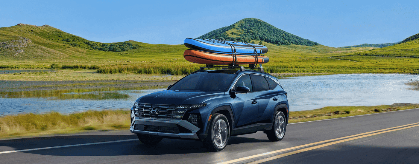 Blue 2026 Hyundai Tucson driving past a lake with paddleboards on the roof.