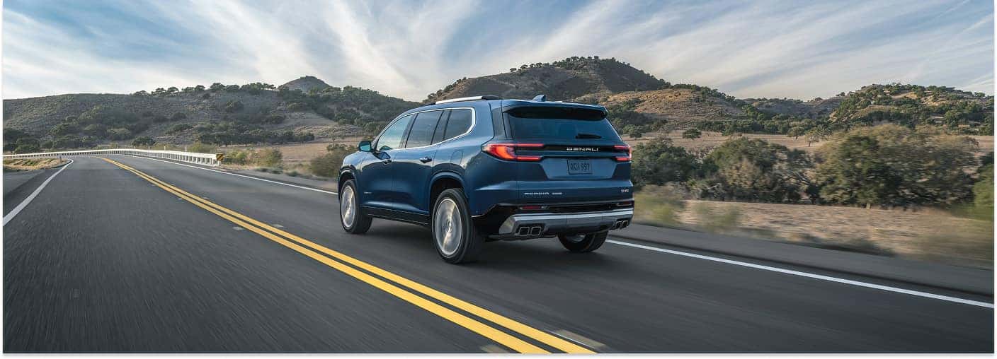 A rear three-quarter view of a blue 2026 GMC Acadia Denali driving along a scenic, two-lane highway bordered by rolling, tree-dotted hills.