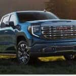 A deep blue 2026 GMC Sierra 1500 Denali pickup truck is parked on a grassy hillside under warm golden sunlight, showcasing its prominent chrome grille and polished multi-spoke wheels.