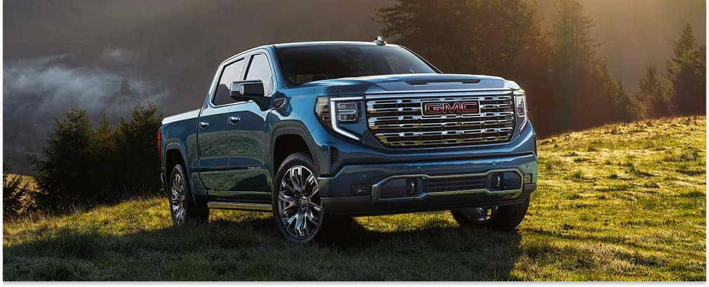 A deep blue 2026 GMC Sierra 1500 Denali pickup truck is parked on a grassy hillside under warm golden sunlight, showcasing its prominent chrome grille and polished multi-spoke wheels.
