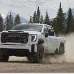 White 2026 GMC Sierra 2500HD pickup truck driving on a dirt road near snowy mountains, front three-quarter view.
