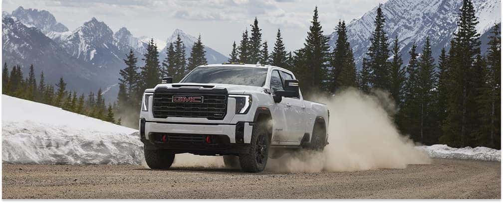 White 2026 GMC Sierra 2500HD pickup truck driving on a dirt road near snowy mountains, front three-quarter view.