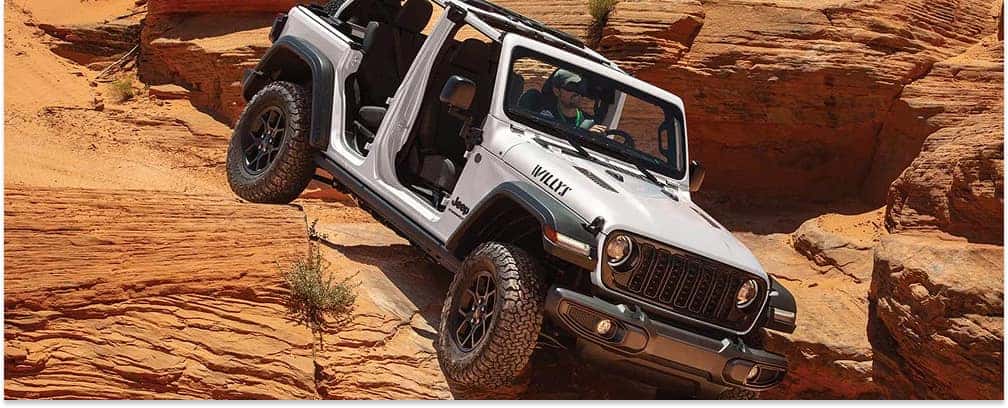 White 2026 Jeep Wrangler Willys with doors off climbing red desert rocks, front three-quarter view.