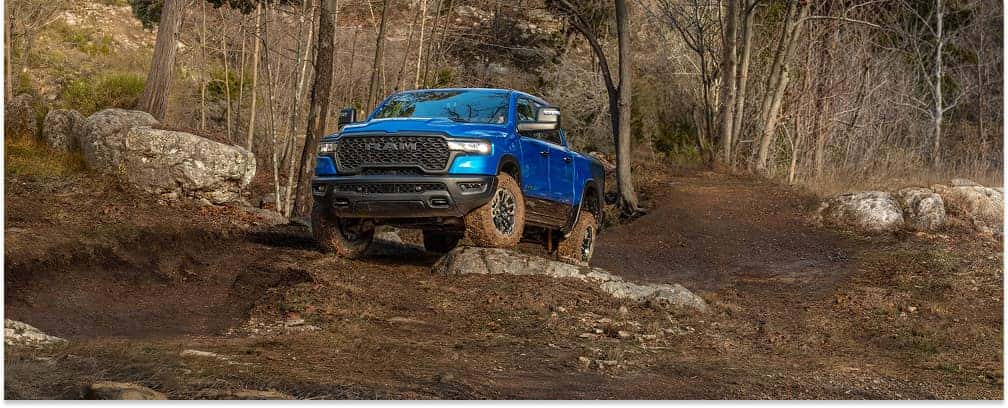 Front three-quarter view of a blue 2026 RAM 1500 Rebel driving over rocky off-road terrain.